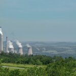 Johnstown, PA, USA - June 12, 2008: Mixed coal and nuclear power plant in Appalachian wilderness under light blue sky. Chimneys and cool towers spew white vapor clouds. Green foliage up front.